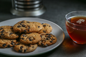 A cup of hot black tea in a glass cup and a teapot press and a gray ceramic plate with cookies with chocolate chips on a gray ceramic tabletop.