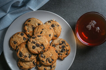 A cup of hot black tea in a glass cup and a gray ceramic plate with cookies with chocolate chips on a gray ceramic counter top with a kitchen towel.