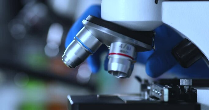 Close up on scientist hand with steriled gloves rotating and adjusting block of microscope lenses. Healthcare technology and pharmaceutical research and development concept.