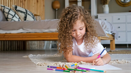 Little preschooler girl with long loose curly hair takes coloured pencil and draws lying on wooden...
