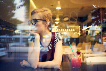 woman with short hair in glasses sits in a cafe at a table with a cocktail relaxing