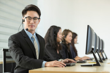 Cheerful Asian man in headset smile with cheerful and happy while working on computer at desk with female coworkers in help desk office. Sevice mind and ready  for marketing support concept