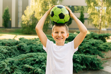 Little boy in white t-shirt holding green soccer ball and smiling at camera. Green background, nature