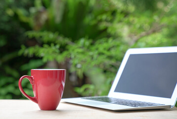 Good morning with coffee in red cup with computer laptop, green garden bokeh background. Copy space.