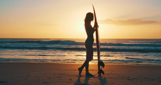 Surfer with board. Slim woman in swimsuit stands on the beach and touches surfing board with waves rolling on the background
