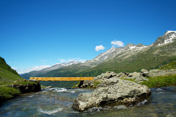 Small wooden bridge over an alpine creek in a meadow with flowers under a deep blue sky