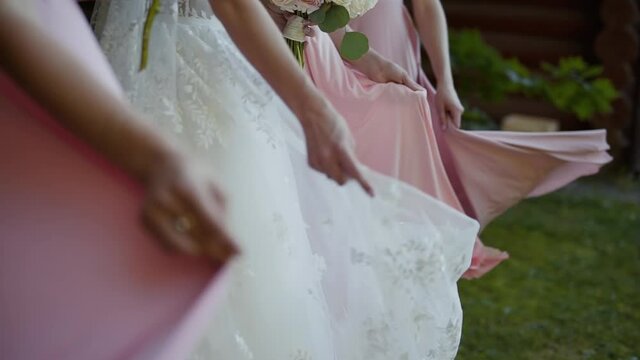 Bride in white dress and bridesmaids in pink dresses with bouquets of flowers are dancing. Woman waving their skirts on the green lawn.