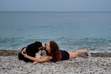 Walking and relaxing with large dog on warm summer morning. Caucasian pretty red haired woman and Bernese Mountain Dog lie together on pebble beach and enjoy each other.