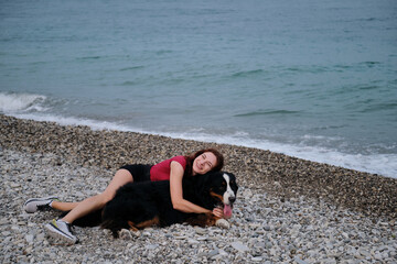 Walking and relaxing with large dog on warm summer morning. Caucasian pretty red haired woman and Bernese Mountain Dog lie together on pebble beach and enjoy each other.
