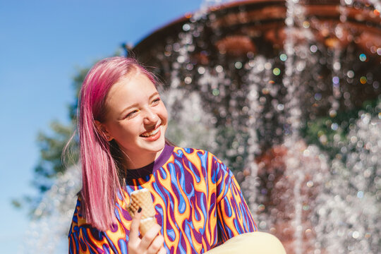 Happy pink-haired teenage hipster girl in a colorful bright T-shirt is eating ice cream on a summer day, with a city fountain in the background.Summer concept.Generation Z style.