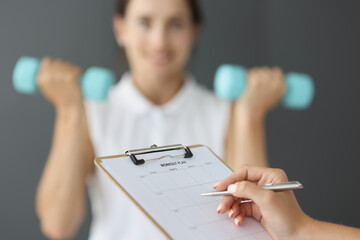 Trainer filling out sports workout plan on background of woman with dumbbells closeup