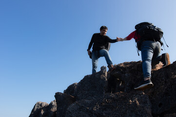 Young man and women backpacker are climbing a mountain, A man stretching his hand to pull a woman up to succeed in their climb, Couple are good teamwork. They both hiked on a clear day.