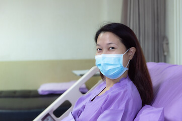 Young Asian female patient is smiling and showing medical mask. Patient feels happy and comfortable with treatment and therapy on hospital bed in hospital room. Medical healthcare concept.