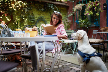 A young beautiful female student is enjoying a company of her dog while reading a book in the bar. Leisure, bar, friendship, outdoor