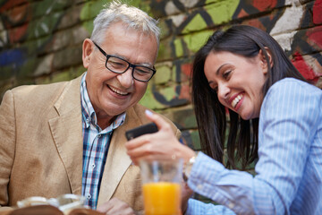 A young beautiful girl is showing a smartphone content to her grandpa while they have a drink in the bar. Leisure, bar, friendship, outdoor