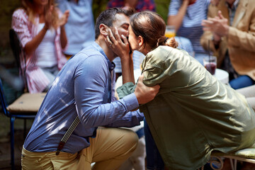 A young man kissing his girlfriend after asking to marry him at a party in a bar with friends. Leisure, bar, love, outdoor