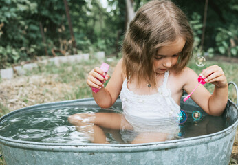 little cute girl in a pink swimsuit blowing soap bubbles and flops in a basin in the summer in the backyard, the concept of a happy childhood and summer vacation