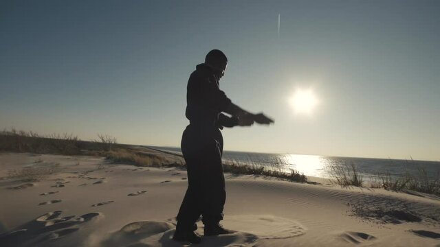 Male dancer turns in sand then drops down on sandy hill by ocean, wide shot slow motion steadicam