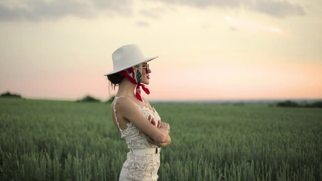 Beautiful girl in a white dress and hat stands in a field at sunset. Girl with painted red lips in a headscarf.