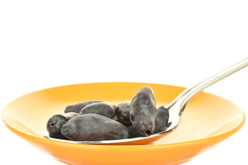 Several berries of dark purple organic honeysuckle with a metal spoon on a ceramic yellow saucer, close-up, isolated on white.