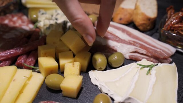 Table with lots of food. Cheese, ham, sausages and olives. A woman's hand takes an olive. Close-up, black background, slow motion, HD.