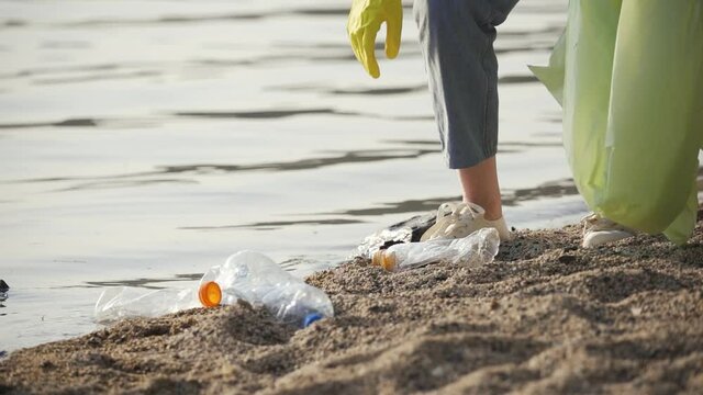 A woman in rubber gloves collects various garbage on the shore of the lake close-up. Preservation of the environment and nature. Slow motion, HD.