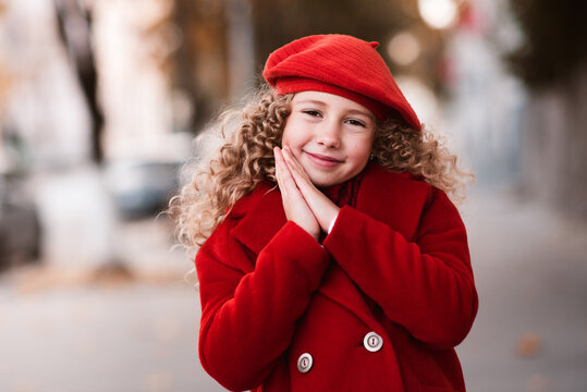 Stylish kid girl 5-6 year old wear red hat and jacket over city street outdoors. Autumn season. Childhood. Happiness. Child with blonde curly hairstyle.