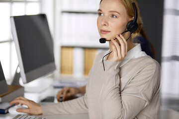 Friendly businesswoman talking by headset while sitting in office. Call center and diverse people group in business
