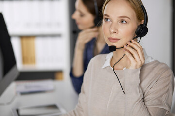 Friendly businesswoman talking by headset while sitting in office. Call center and diverse people...