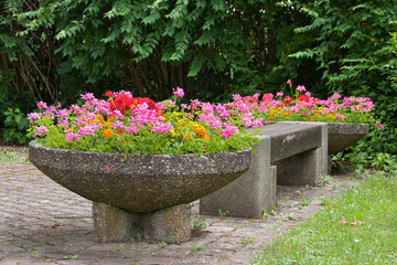 Eine Siitzbank aus Beton mit zwei mit bunten Blumen bepflanzten Betonkübeln an den Stirnseiten