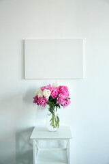 Canvas mockup hanging on white wall and vase with pink flowers on wooden shelf