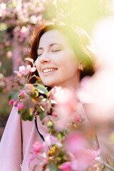 Fototapeta premium Close-up portrait of a young girl with a smile on her face standing around a blooming rose tree in the garden. The concept of spring and allergy to blossom.