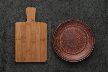 Brown wooden cutting board, empty brown clay plate on black stone table. Top view with copy space.. Rustic dishes