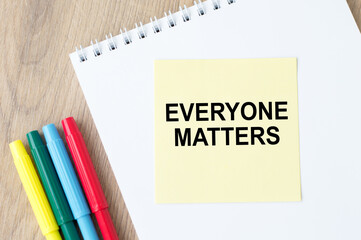 Text EVERYONE MATTERS on a yellow card that lies on a white notepad next to colored markers on a wooden table