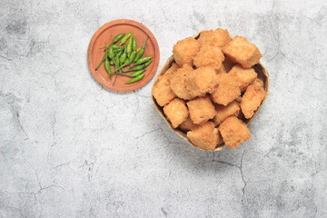 Photo Flat Lay, tahu krispi or crispy Tofu  and green chili at bamboo bowl, besek under brown abstract marble 
