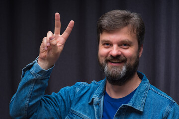 A positive young man smiles. The raised hand symbolizes the victory sign. A stylish beard and hair with a noble gray streak. The background is blurry gray. Space for text