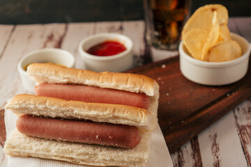 High view of a pair of plain hotdogs with pots of mustard and ketchup and some potato chips on a rustic wooden tray. Horizontal view. Fast and junk food concept.