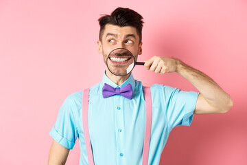 Funny man in bow-tie smiling, showing teeth with magnifying glass and looking aside at logo,...