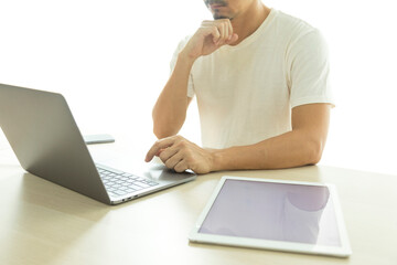 Young man in white shirt sitting in front a computer laptop and tablet on wooden working desk in living room.