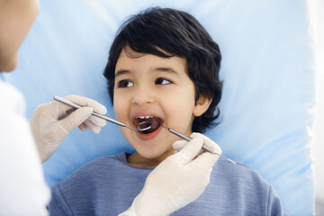 Cute arab boy sitting at dental chair with open mouth during oral checking up with doctor. Visiting dentist office. Stomatology concept