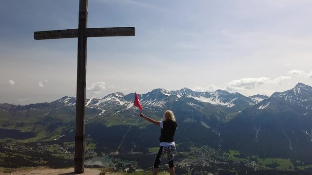 SLOW MOTION: Woman with Swiss flag on Piz Scalottas cross. Tourist resort for ski activities in winter and bike in summer. Plessur Alps of Switzerland in Grisons.
