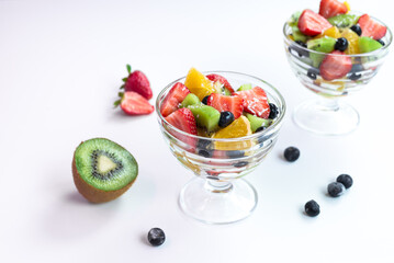 pieces of fresh fruits and berries with whipped cream and chocolate in a transparent bowl on a white table. 