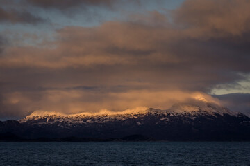 Naklejka na ściany i meble Morning sun over clouds flying over Andes mountain peaks in a little island in Ushuaia, Tierra del Fuego, Argentina. Waters are of the Beagle Channel. 