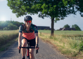 Frontal photo of a young woman cyclist, smiling, riding her road bike, in the middle of nature, illuminated by sunlight. She is wearing a helmet, sunglasses, and a pink cycling outfit.