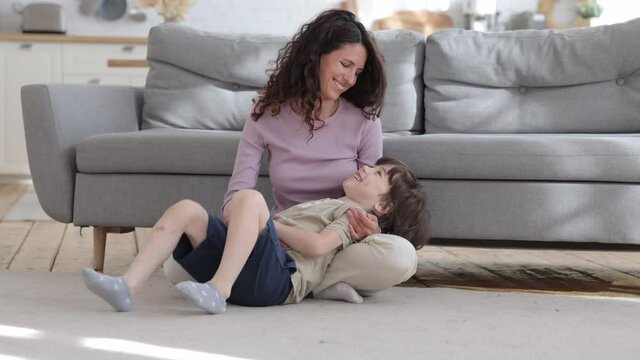 Overjoyed loving mom has fun with small son tickling boy sitting with kid on floor carpet in living room laughing. Cheerful caucasian woman mom or baby sitter enjoy funny playing with child at home