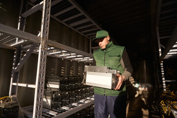 Mover holding bitcoin ASIC miner in warehouse. Worker with ASIC mining equipment on stand racks for...