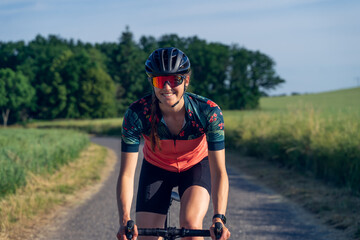 Naklejka premium Motion Portrait of a young woman cyclist, smiling, riding her road bike, in the middle of nature, illuminated by sunlight. She is wearing a helmet, sunglasses, and a pink cycling outfit.