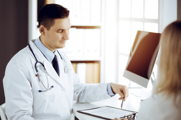 Fototapeta premium Friendly male doctor and patient woman discussing current health examination while sitting in sunny clinic. Perfect medical service in hospital