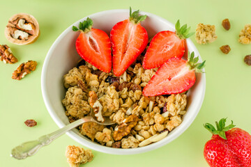 Healthy breakfast in a white bowl with natural granola. Homemade baked muesli, nuts, fresh strawberries on a green background.