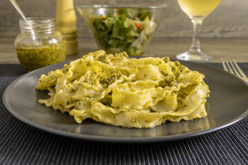 Pasta Mafaldine with tofu pesto sauce served on a dark plate. Salad bowl, glass jar with pesto sauce, a glass of wine and pepper mill. Textile placemat, wooden kitchen surface.